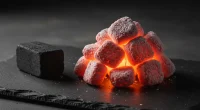 Контрастное изображение горящих кокосовых углей и одного необработанного древесного угля на тёмном фоне.